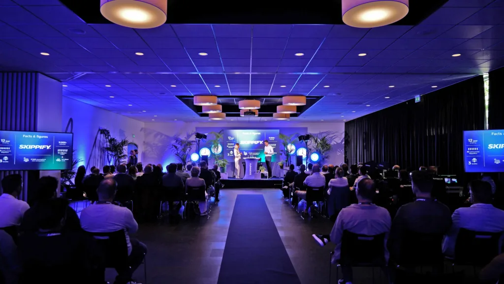Overzicht van de volle evenementzaal in Walibi Holland tijdens de merkreveal van Skippify, met publiek dat luistert naar de presentatie op het blauw verlichte podium